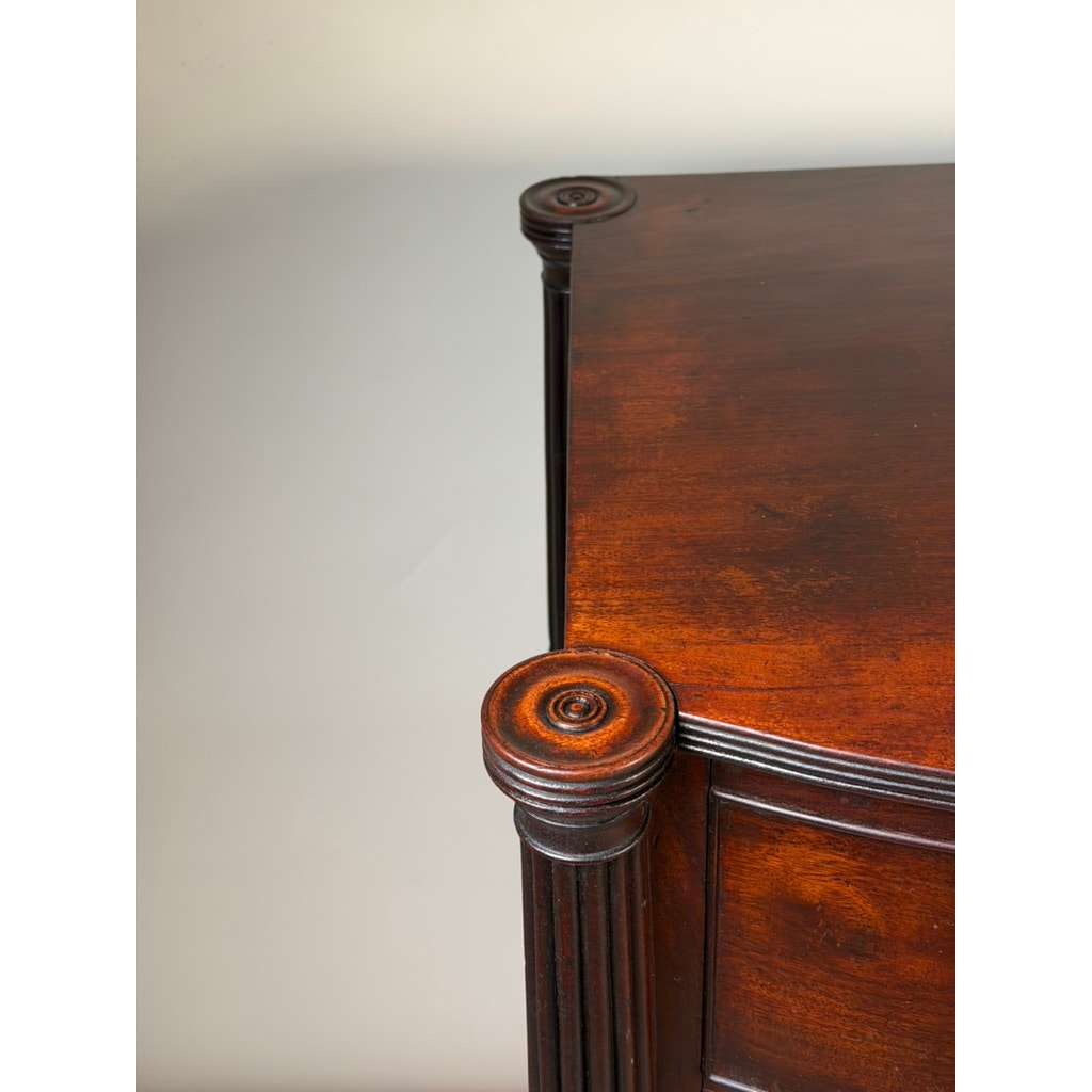 Close-up of a polished corner of an Early 19th Century New England Federal Mahogany Bow-Front Chest of Drawers (c. 1810–1825), featuring carved circular decorative details and vertical grooves on the posts, set against a plain light background.