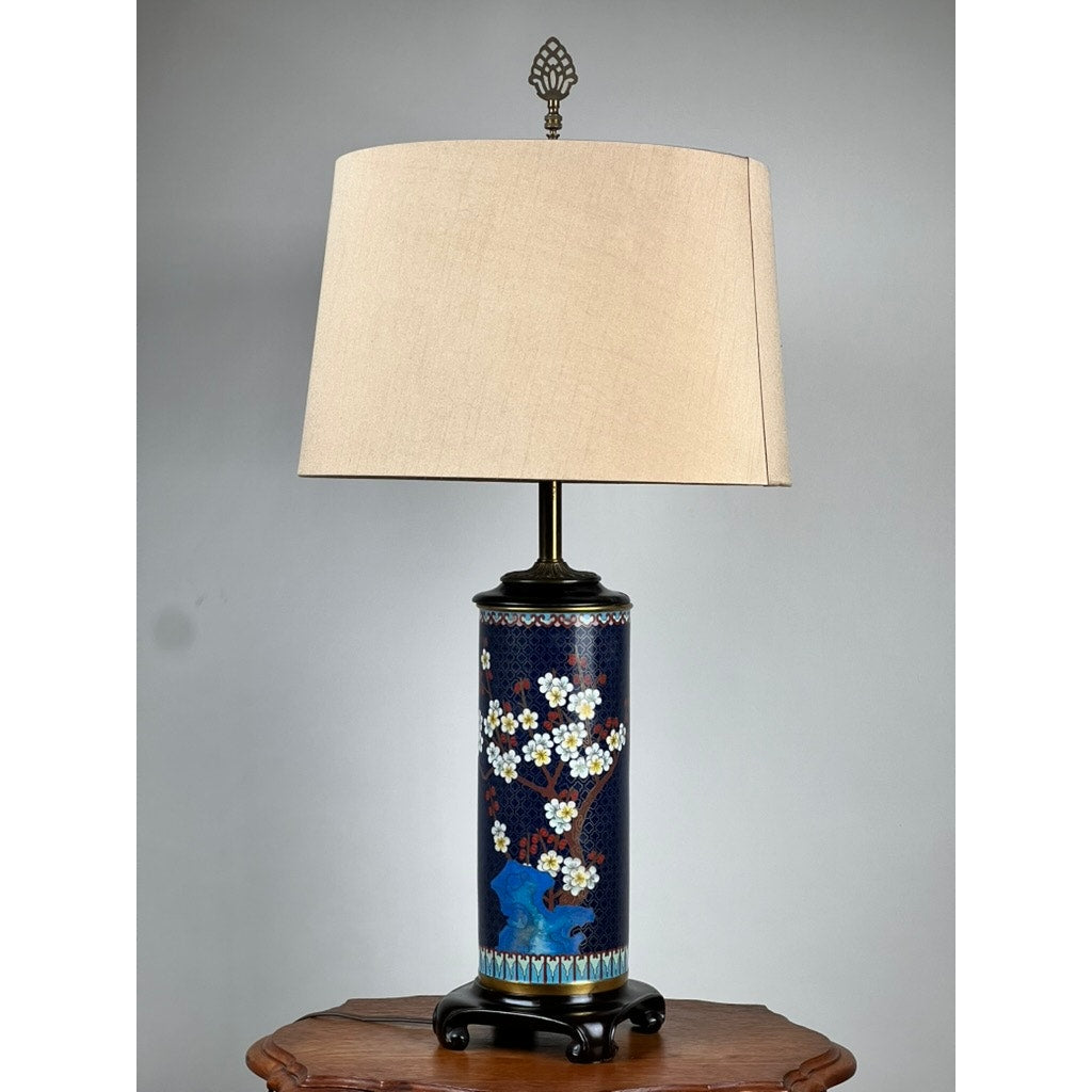 The Vintage Japanese Cloisonné Cylinder Table Lamp by Knob Creek features a blue cylindrical base with floral and bird designs, a beige shade, and sits on a wooden table against a neutral wall.