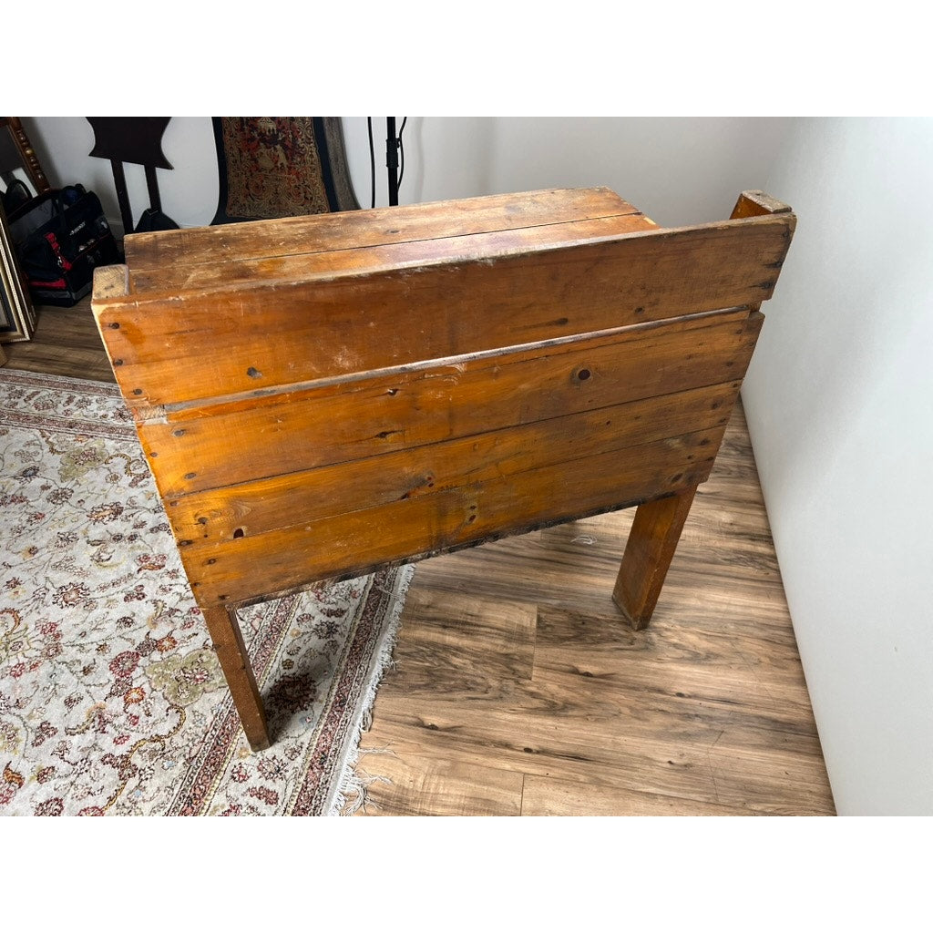 A rustic wooden table on a wood floor near a white wall and patterned rug is accompanied by a Red Fox Antique Tin Lined Pine General Store Ginger Ale Cooler from Rhode Island, c. 1930, with additional decor in the background.