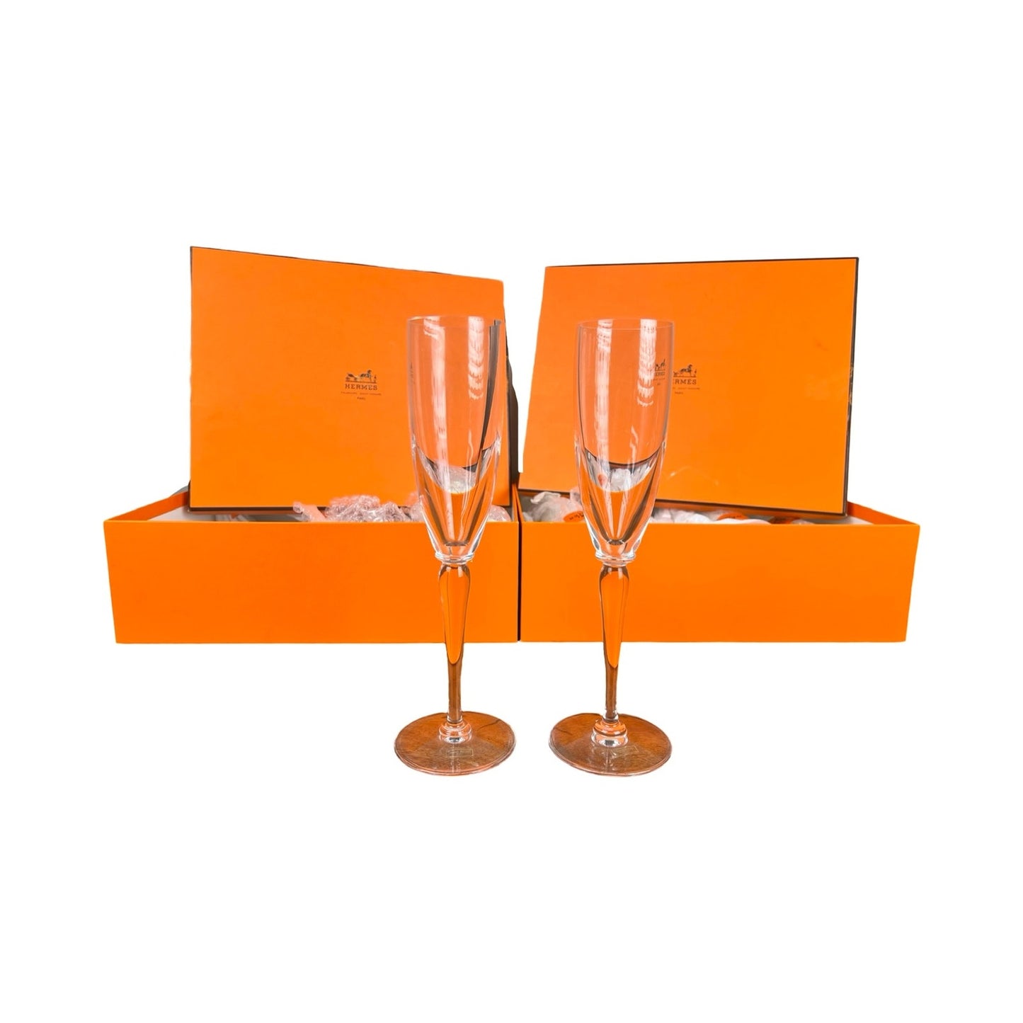 A set of eight Hermès crystal champagne flutes by Saint Louis France (10.5") is displayed in front of two orange Hermès boxes with open lids and packaging, all set against a white background highlighting this luxury glassware.