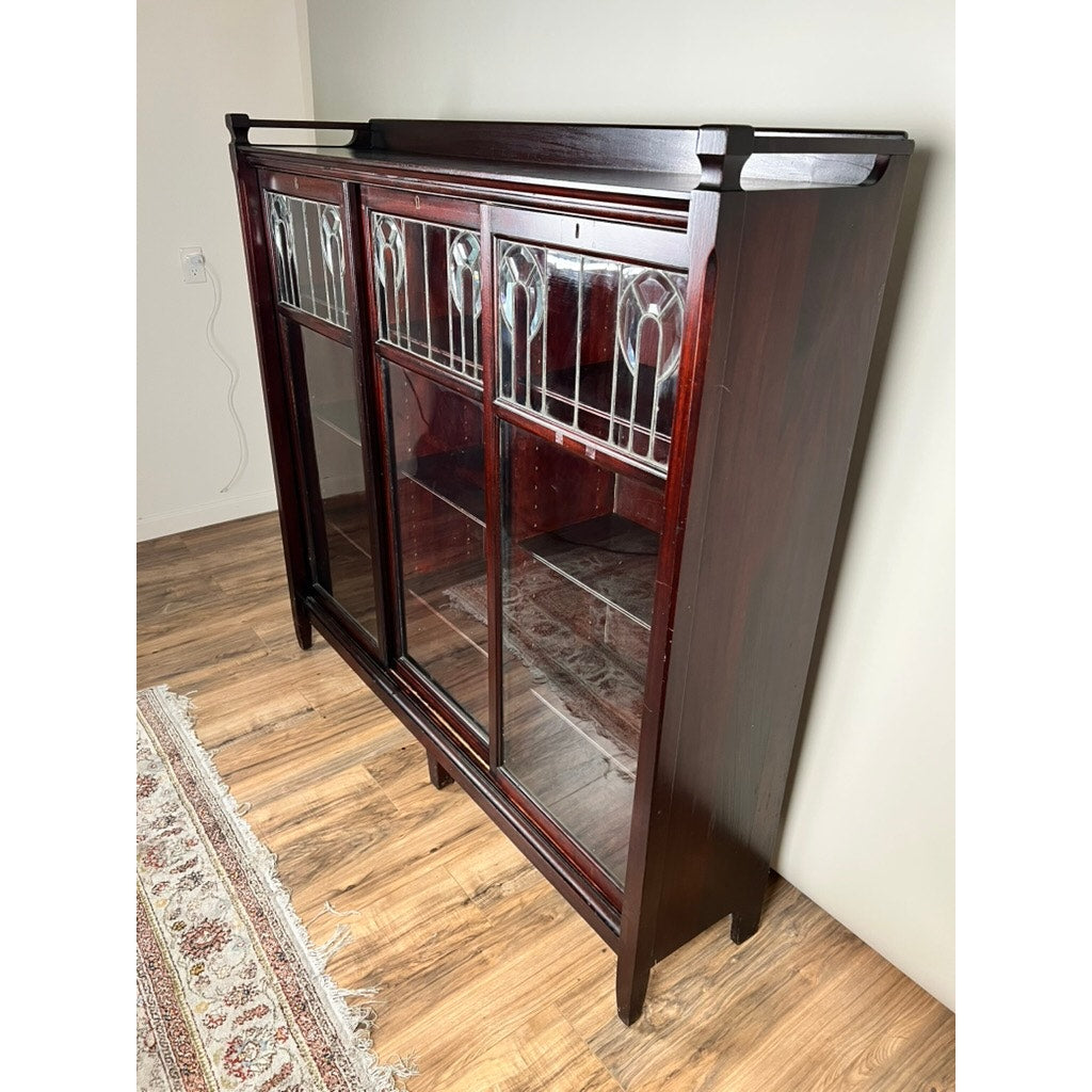 An Antique Arts & Crafts Leaded Glass Mahogany Bookcase c. 1920 stands on a wooden floor by a white wall and ornate rug, showcasing three sections with visible shelving and decorative glass panel doors.