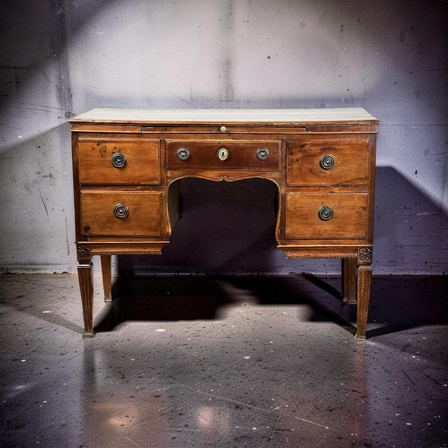 An Antique Louis XVI Period Ladies Desk with a tooled leather top from the 18th century stands on a concrete floor against a worn wall, its five drawers and ornate metal handles highlighted by dramatic lighting. Brand unknown.