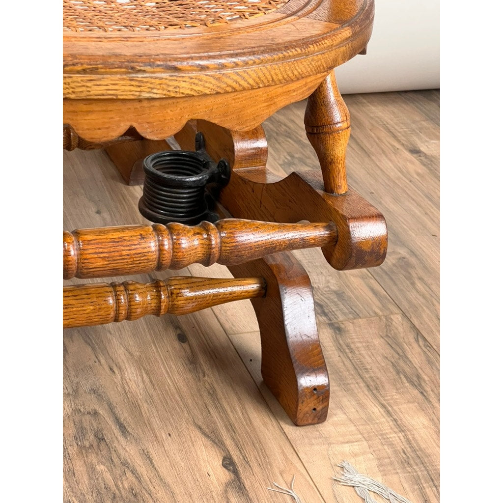 Close-up of the finely turned wooden leg and support of an Antique Victorian Oak Platform Rocker (c. Late 19th Century), highlighting its woodwork and the exposed black metal spring beneath the seat on a wood floor. Brand: Unknown.