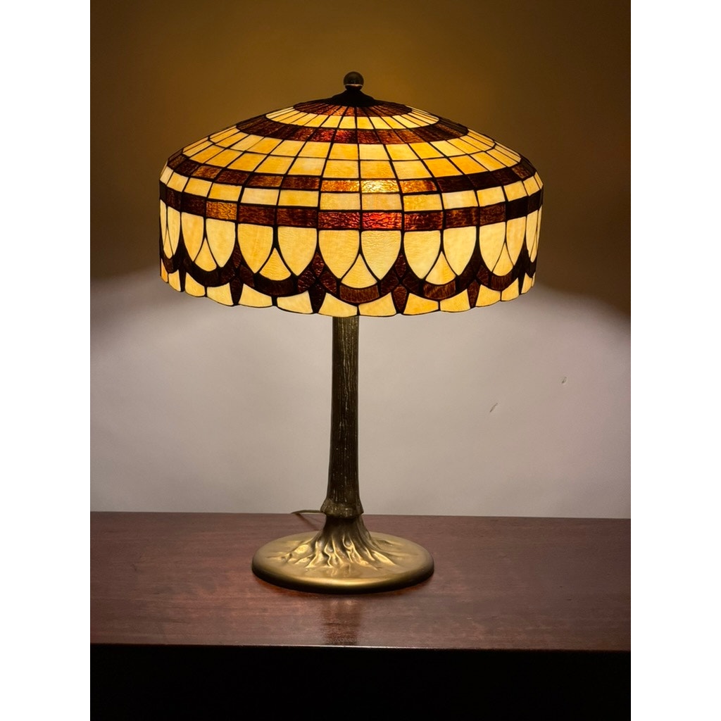 A Pearl City Antiques Vintage Tiffany Revival Leaded Stained Glass Table Lamp with a cast brass tree-form base sits on a wooden surface, its yellow and brown geometric-patterned shade casting a warm light on the wall behind it and adding an elegant touch to the room.