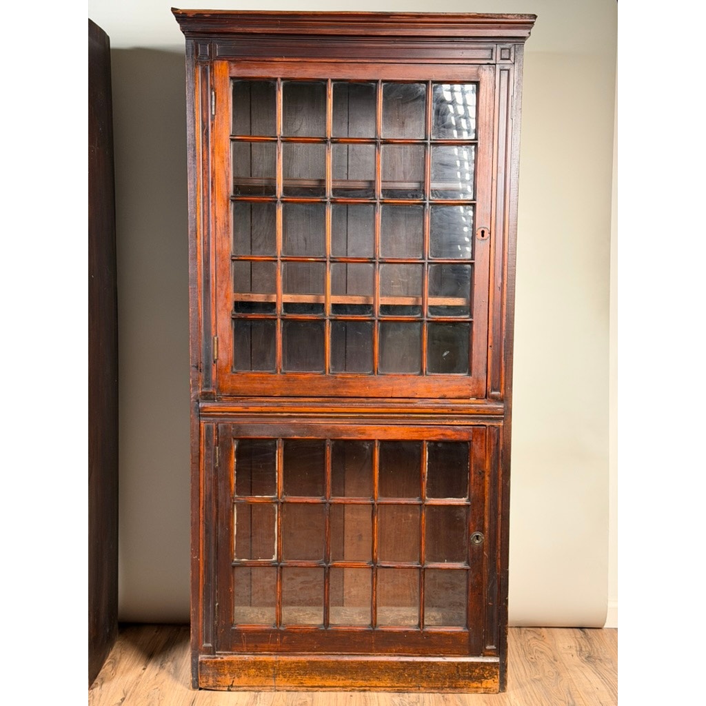 Standing on a wooden floor against a light-colored wall, this pair of mid-19th century New England cherry glazed display cupboards from Pearl City Antiques (c. 1840–1860) features tall wooden construction with two glass-paned doors—one upper and one lower—each sectioned into squares, reminiscent of early American cabinetry.