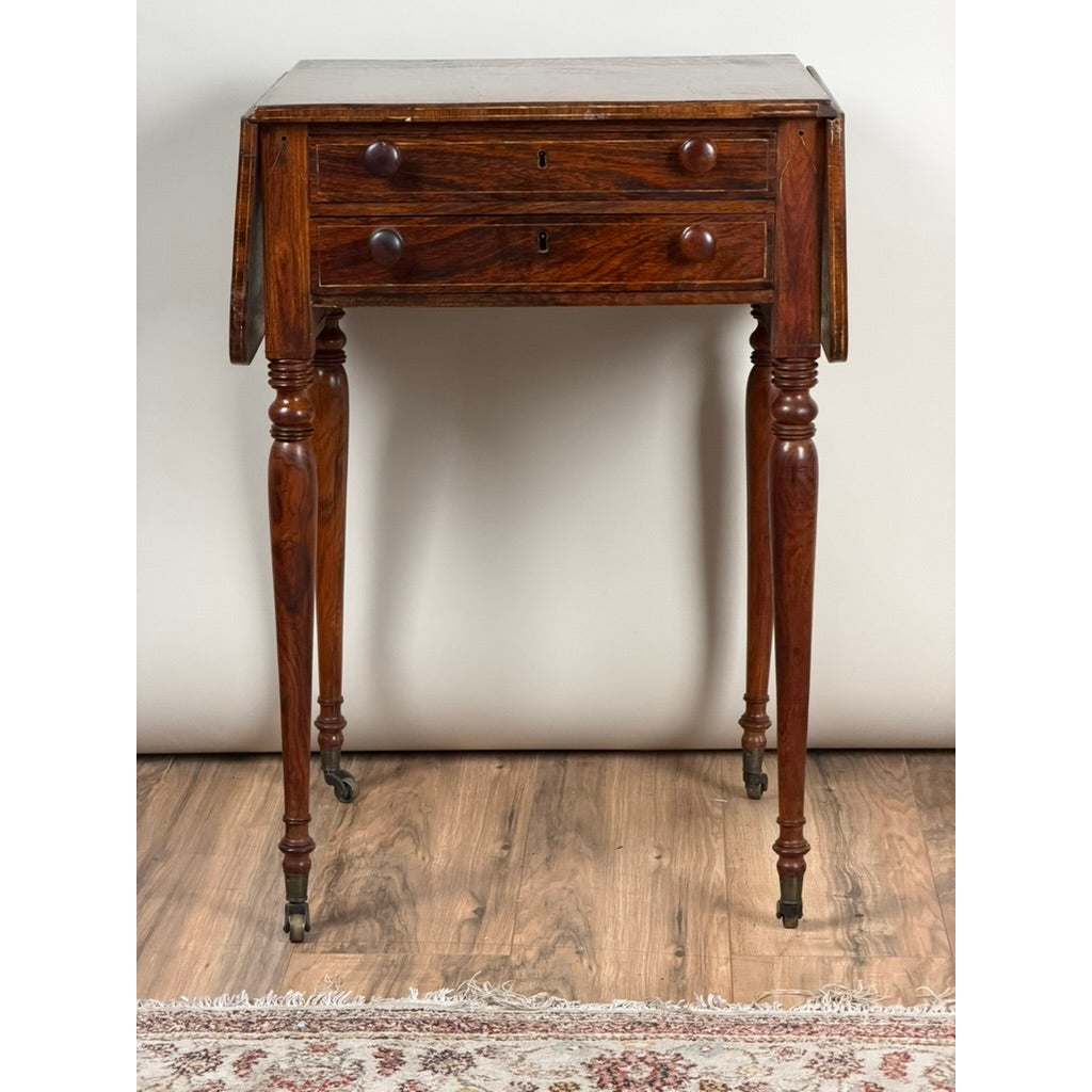 An English Regency Rosewood Two-Drawer Drop-Leaf Work Table with Brass and Satinwood Inlay, featuring dark wood, two drawers, turned legs, and metal casters.