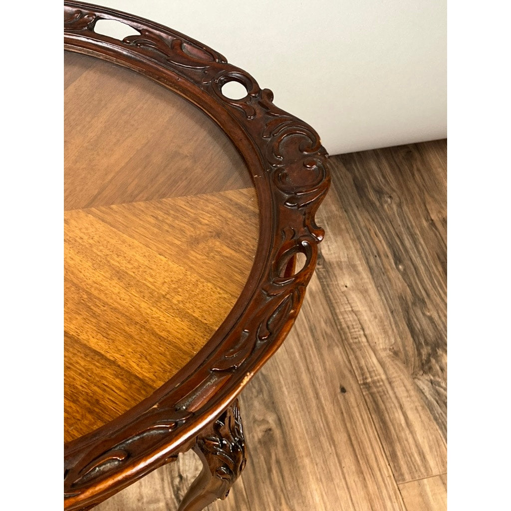 A Vintage Louis XVI Style Butlers Table with Removable Glass Tray is shown close up, featuring ornate carved edges. The round wooden table stands on a wood-patterned floor beside a light-colored wall. Brand unknown.
