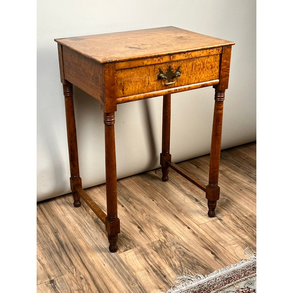 An Antique Sheraton Birdseye Maple One Drawer Stand (c. Early 19th Century) with a single ornate-handled drawer and turned legs with stretchers sits on a wood-patterned floor near a rug. Brand: None/Unknown.