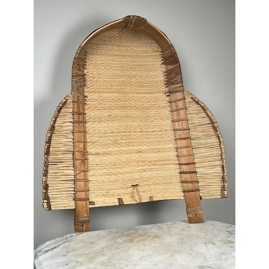 The Vintage Traditional Winnowing Basket, made from natural fibers with curved edges and vertical wooden supports, stands upright on a light marble surface against a plain gray wall. Brand unknown.