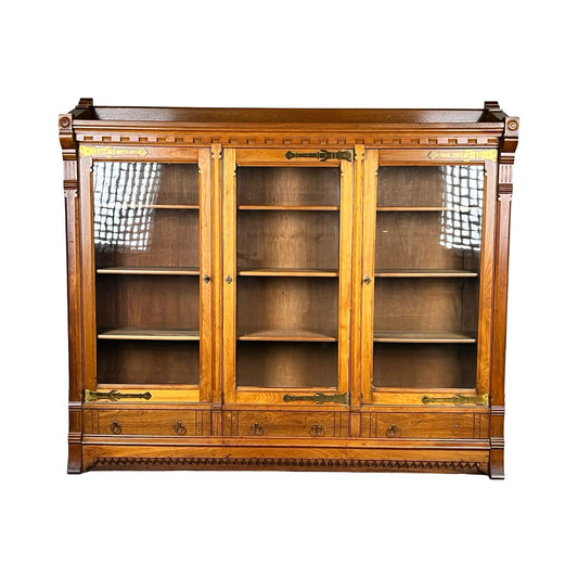 Antique Eastlake Victorian Walnut Triple Bookcase c. 1880 with three glass doors, brass accents, four lower drawers, decorative carvings, and multiple shelves for storage. Brand: Unknown. Set against a white background.