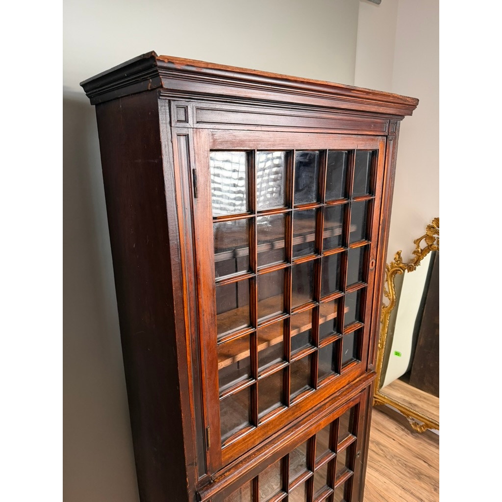 A pair of mid-19th century New England cherry glazed display cupboards (c. 1840–1860) from Pearl City Antiques stand in a room with light walls and a wooden floor, their glass-paned doors arranged in a grid pattern reminiscent of early American cupboards and accented with decorative trim. Nearby, a large ornate gold-framed mirror completes the elegant setting.
