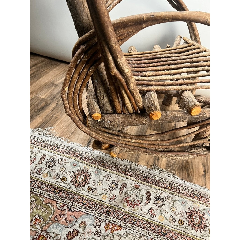 Close-up of a Vintage Adirondack Bent Willow Arm Chair crafted from natural branches, placed beside a decorative patterned rug on a wooden floor. The photo emphasizes the chair’s rustic texture and the intricate rug design. Brand: None/Unknown.