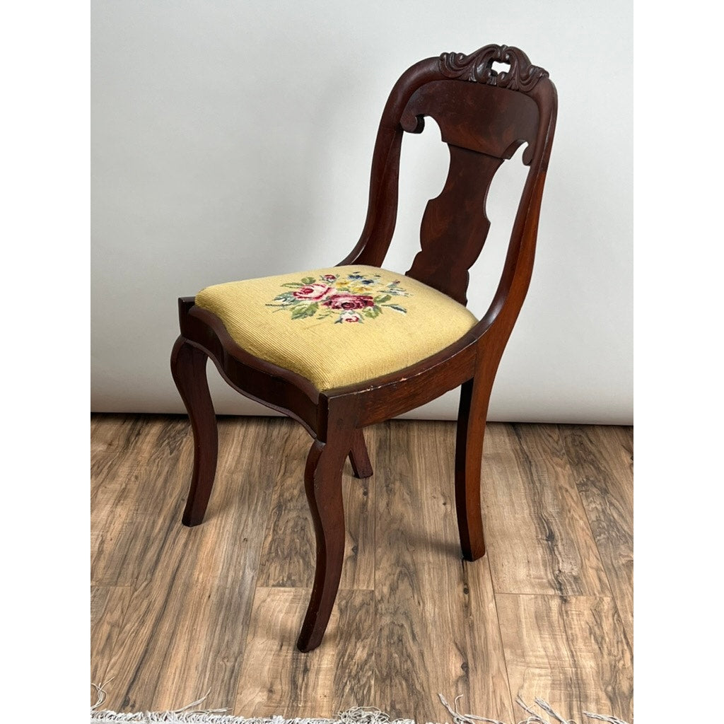 An Antique American Empire Flame Mahogany Saber Leg Side Chair (mid-19th century) with a carved backrest and yellow floral needlepoint upholstery is displayed on a wooden floor against a plain white background.