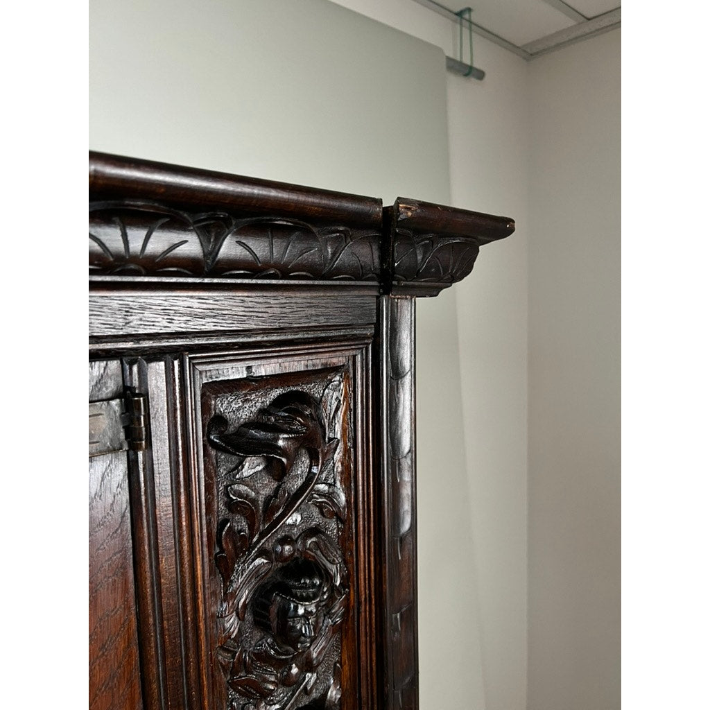 Close-up of the top corner of an Antique 19th-Century French Breton-Style Carved Oak Cabinet featuring deeply carved knights, ornate carvings, and decorative trim, set against a light-colored wall and partially visible ceiling.