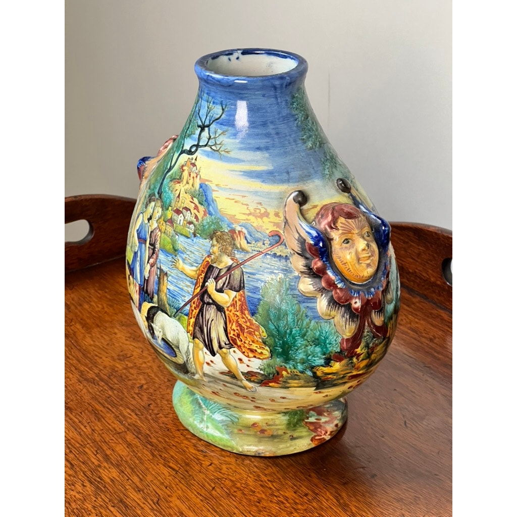 An Antique Italian Renaissance Revival Hand Painted Maiolica Vase with winged putti handles (c. 1920–1940) sits on a wooden surface, featuring colorful Renaissance-style scenes and classical figures against a neutral background.