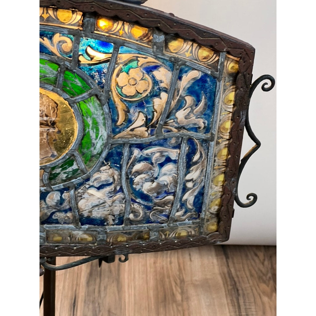 A close-up of an Antique Five Burner Lucerne Whale Oil Lamp with a Napoleon I Fireplace Screen Shade, featuring intricate blue, green, and yellow stained glass and ornate metal framing, displayed on a stand over a wooden floor.
