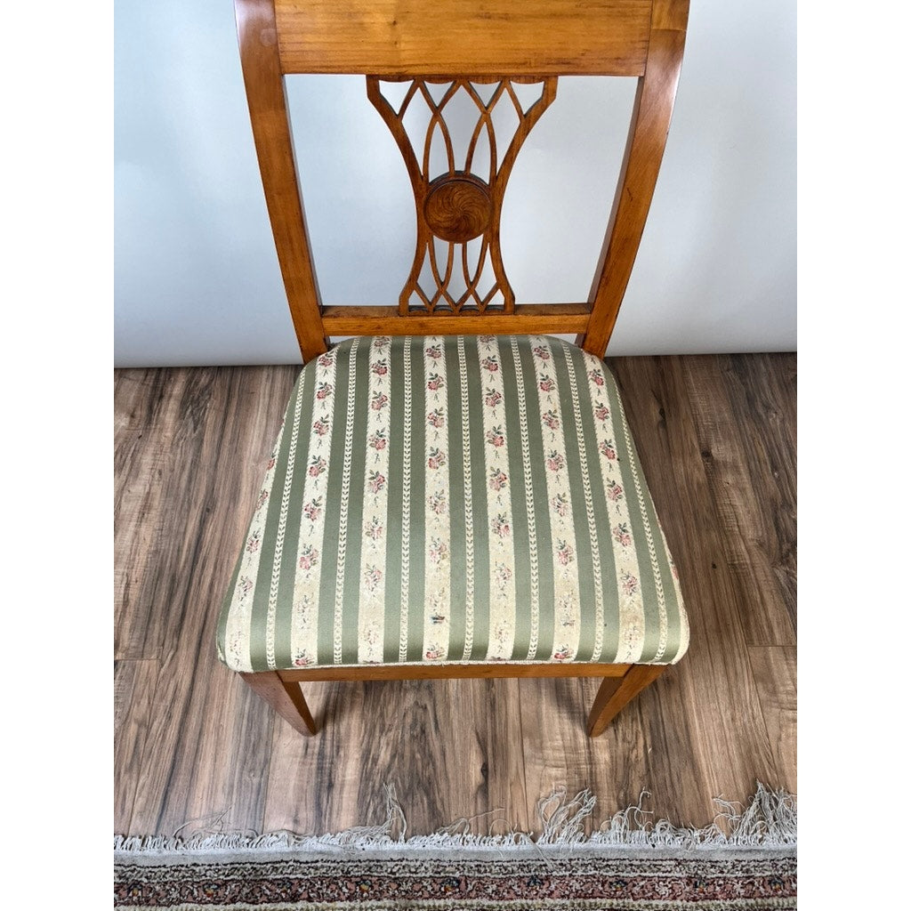 An Antique German Biedermeier Style Cherry Side Chair (early 20th century) with an ornate carved backrest and green-beige striped floral upholstery enhances the elegance of any room, shown beside a fringed rug on a wood floor.