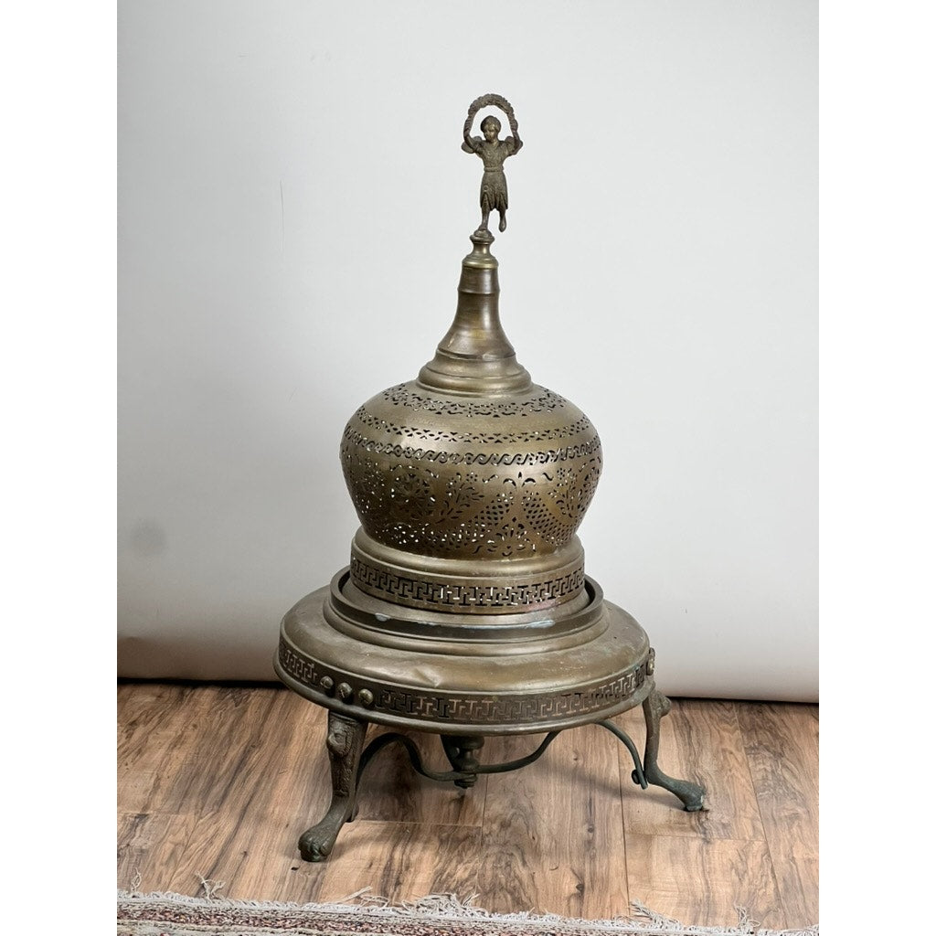 An Antique Venetian Brass Bell Room Heater Brazier (19th Century), featuring ornate cutout patterns, a domed lid with finial, and three curved legs, serves as an elegant decorative piece on the wooden floor.
