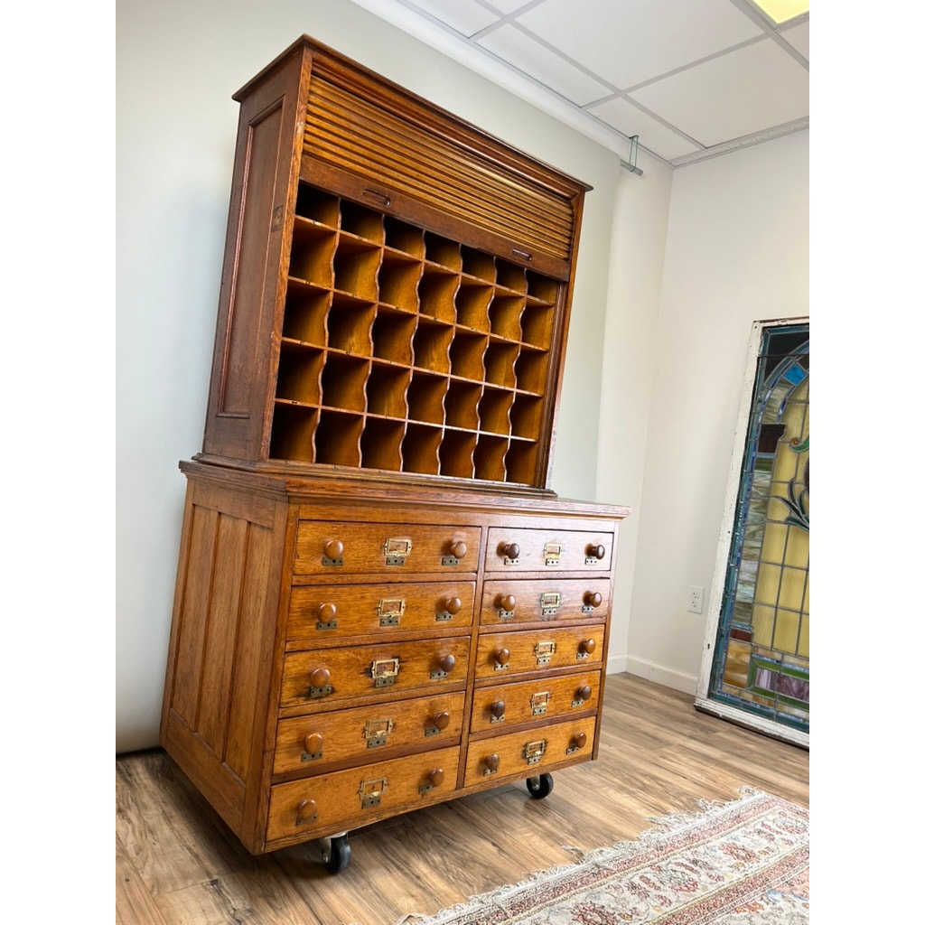 An Antique Early 20th Century Oak Multidrawer File with Roll Top Pigeon Hole Top stands on caster wheels in a room with wooden floors, accompanied by a stained glass panel leaning against the wall.