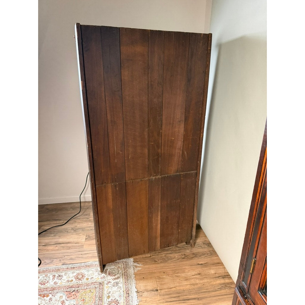 A pair of mid-19th century New England cherry glazed display cupboards by Pearl City Antiques, dating from circa 1840–1860, stand with their backs facing the camera, showcasing vertical dark cherry wood panels reminiscent of early American cabinetry. The cupboards are positioned on a wooden floor near a beige wall and a patterned rug.