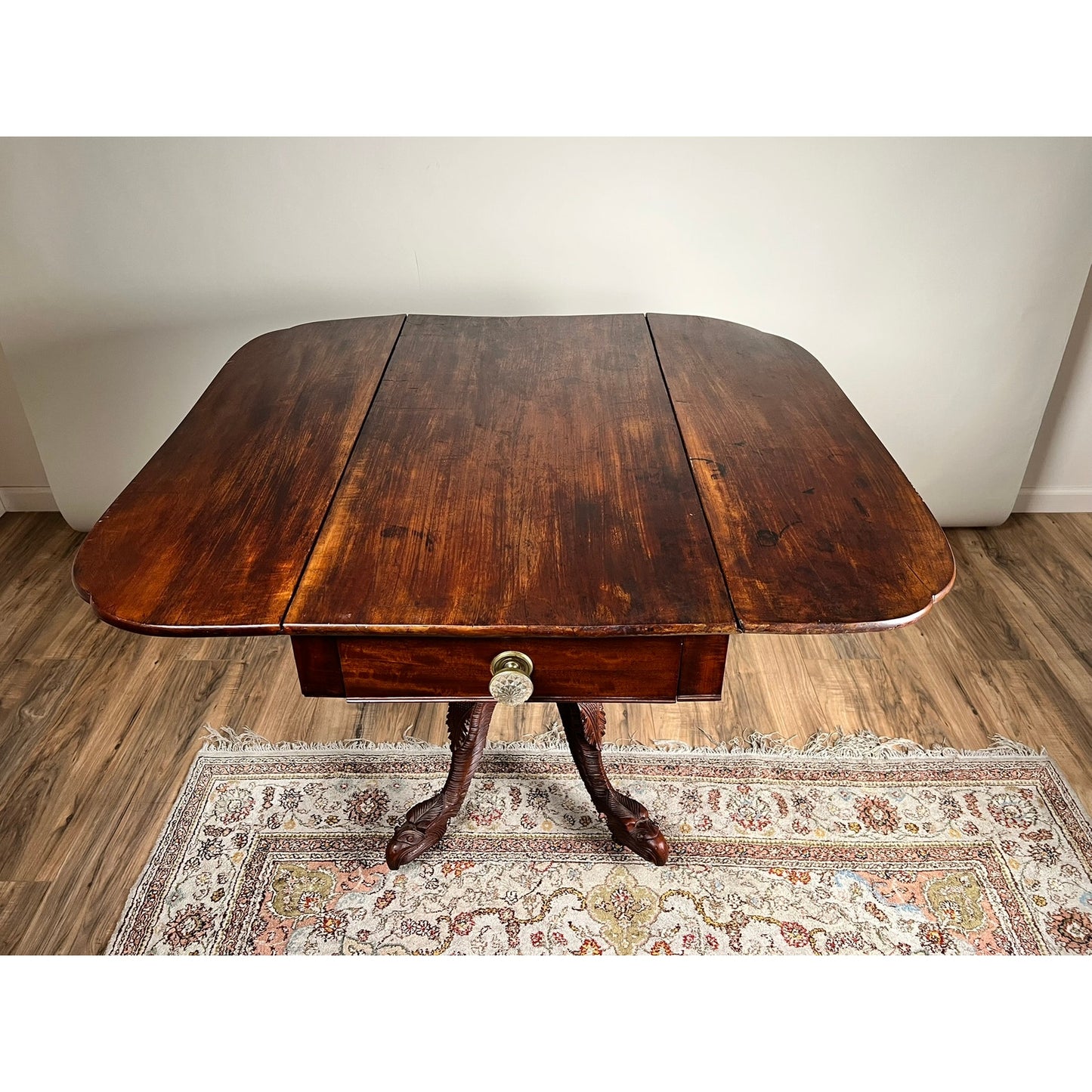 An Antique American Empire Drop Leaf Table c. 1840 with striking eagle-carved feet stands on a decorative rug atop a wood floor, set against a plain light wall. Brand: None/Unknown.