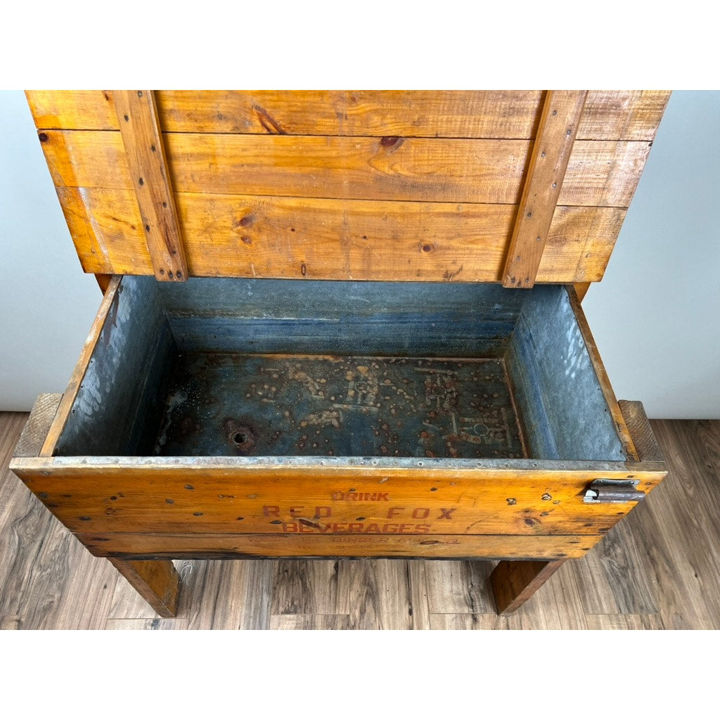An Antique Tin Lined Pine Red Fox Ginger Ale of Rhode Island Cooler c. 1930 sits on a wooden floor, its open lid revealing a metal-lined interior and faded exterior lettering from the historic Red Fox brand.