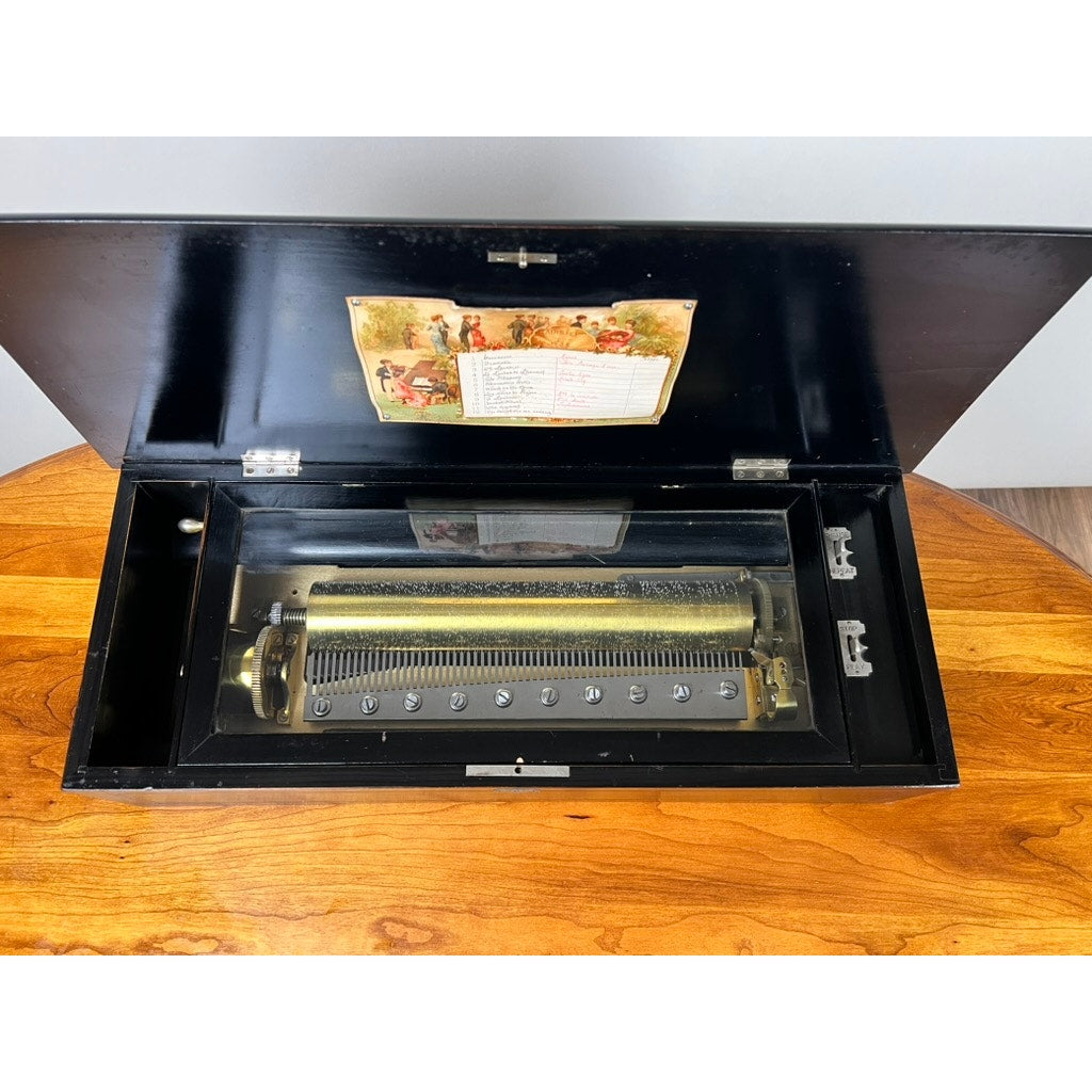 The Antique Swiss 12 Tune Music Box (late 19th century) features an open black wooden case with a glass lid, revealing its brass mechanism inside. A colorful illustration and label adorn the lid, capturing classic 19th-century charm. Brand unknown.