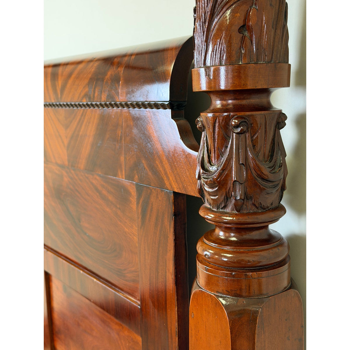 Close-up of an American Classical Carved Mahogany Four-Poster Tester Bed, possibly New York, circa 1825–1840, showing a carved post with acanthus leaf motifs connected to a curved, polished mahogany headboard.