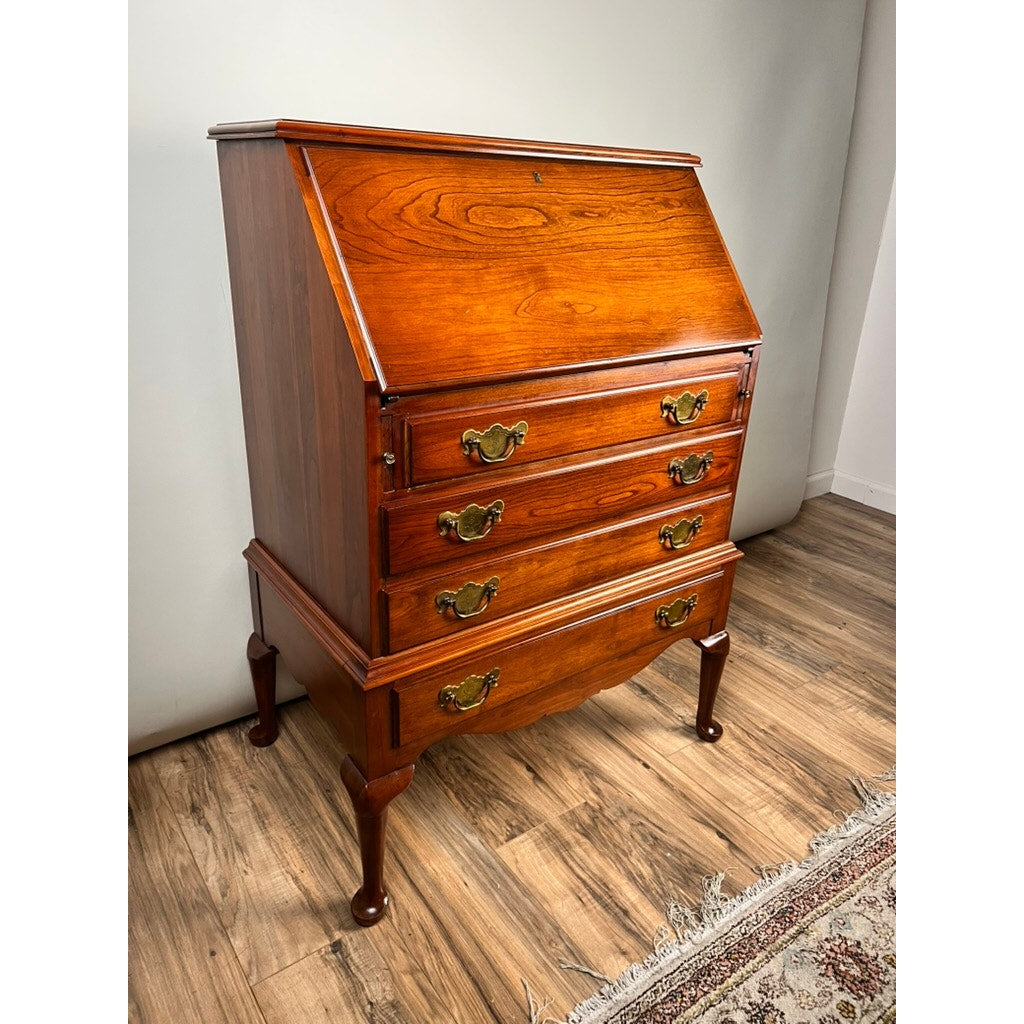 A Vintage Solid Cherry Queen Anne Style Slant Front Secretary Desk by Pennsylvania House with three brass-handled drawers stands on a wood floor beside a patterned rug and plain white wall.