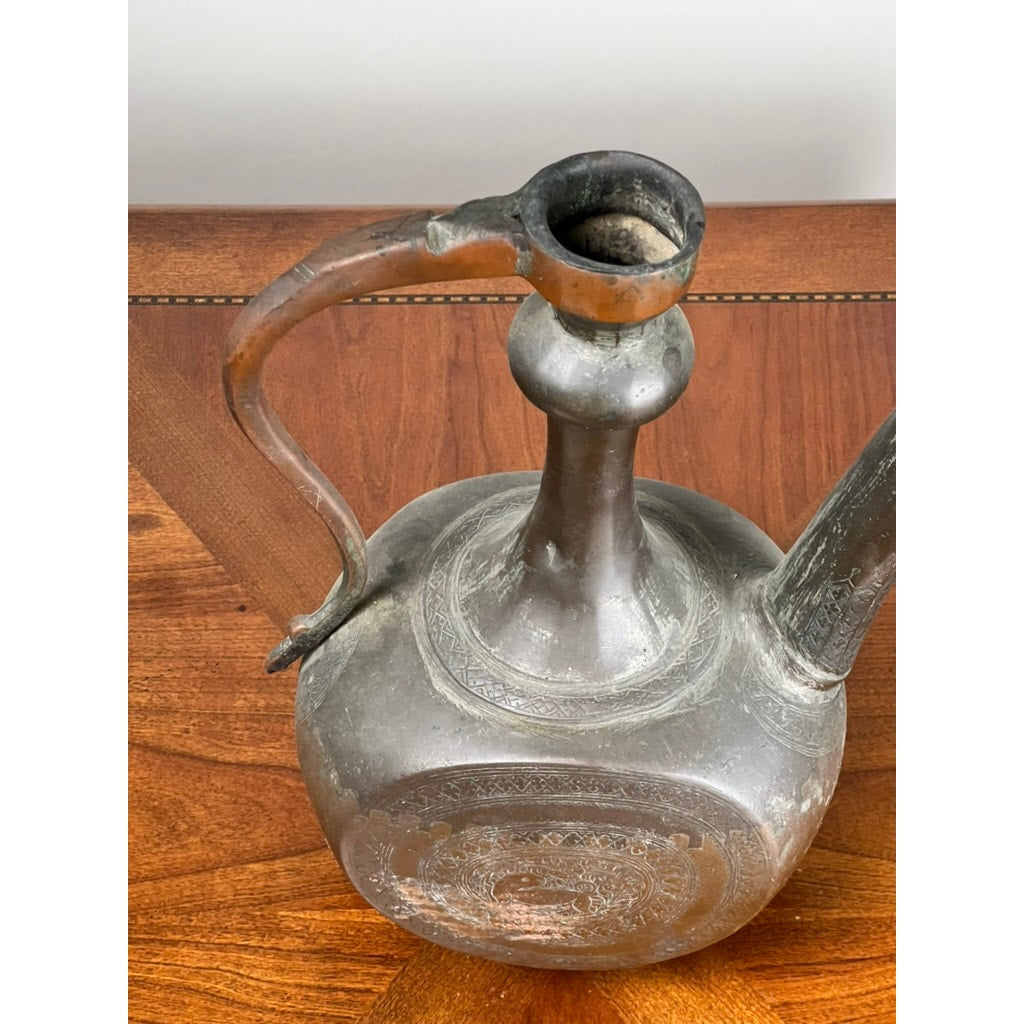 A top-down view of an Antique Qajar Persian Copper Engraved Ewer (19th Century) with curved handle, narrow neck, and spout, displayed on a wooden surface with patterned inlay, highlights exquisite Persian metalwork. Brand: Unknown.