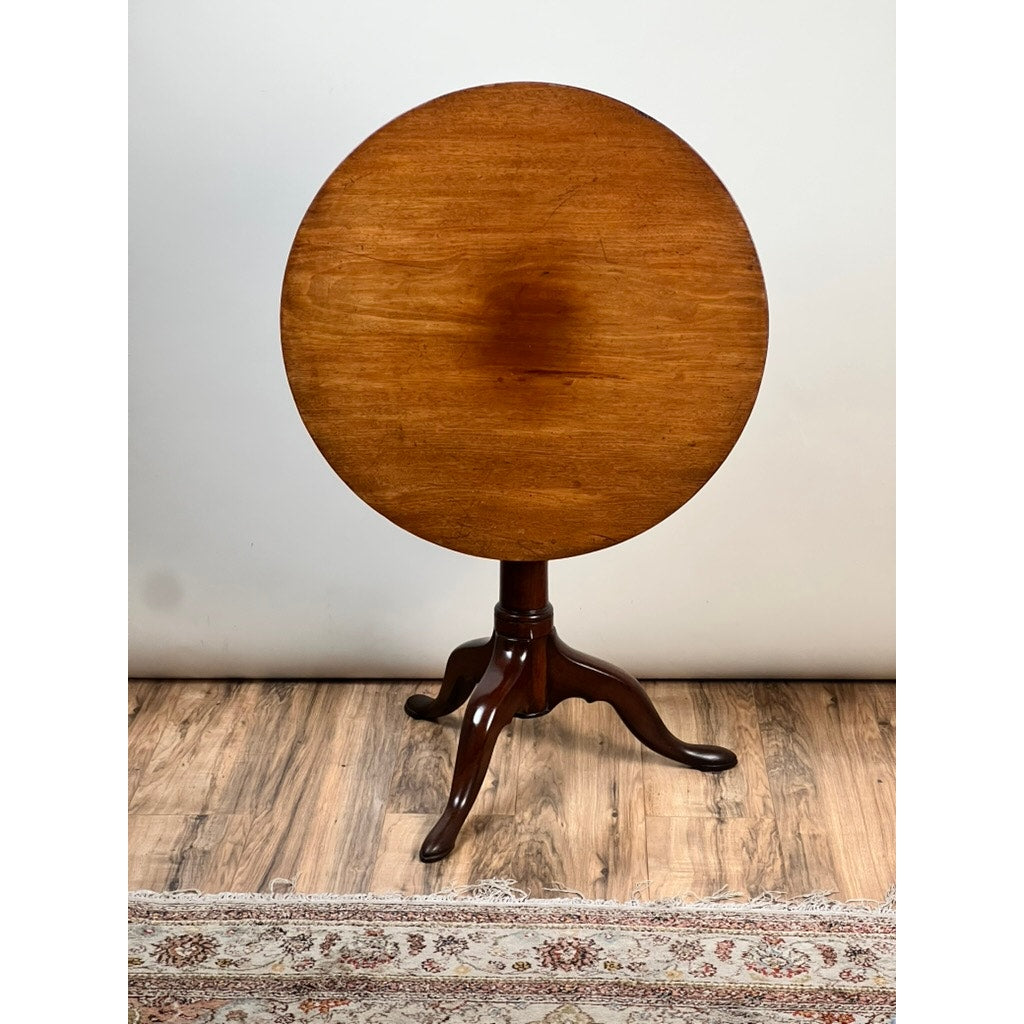 An Antique Queen Anne Mahogany Birdcage Tilt Top Breakfast Table (early 19th century) with a polished brown surface and three curved legs stands on a wooden floor by a patterned rug, against a plain light-colored wall.