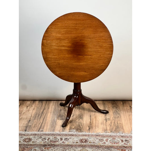 An Antique Queen Anne Mahogany Birdcage Tilt Top Breakfast Table by Pearl City Antiques, dating from the early 19th century, featuring a polished round tilt-top surface and three graceful curved legs, stands on a wood floor beside a patterned rug against a plain light wall.