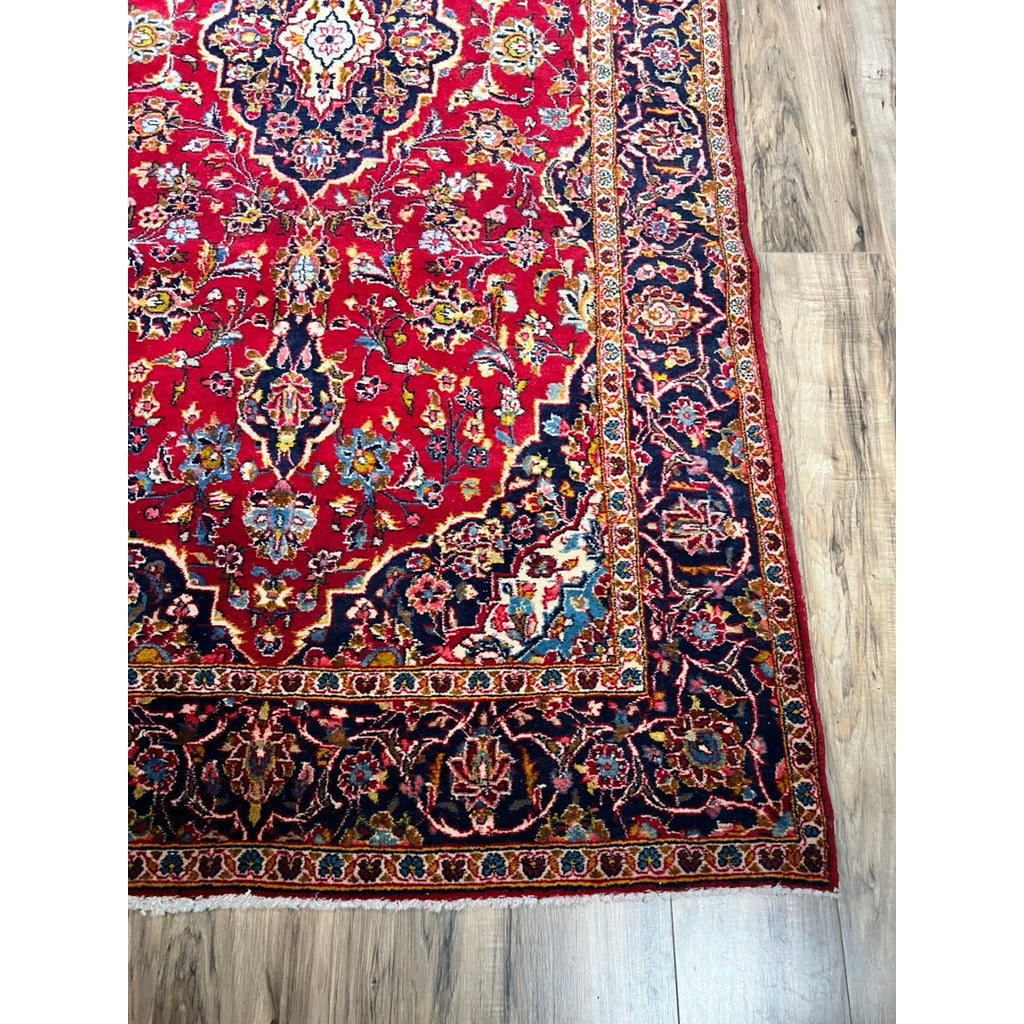 A corner of a vintage hand-knotted Persian Kashan wool gallery rug (c. 1950–1970, 4’5 x 8’8) with intricate red, navy, and cream floral patterns, detailed borders, and fringe is shown on a light wood floor. Brand: unknown.
