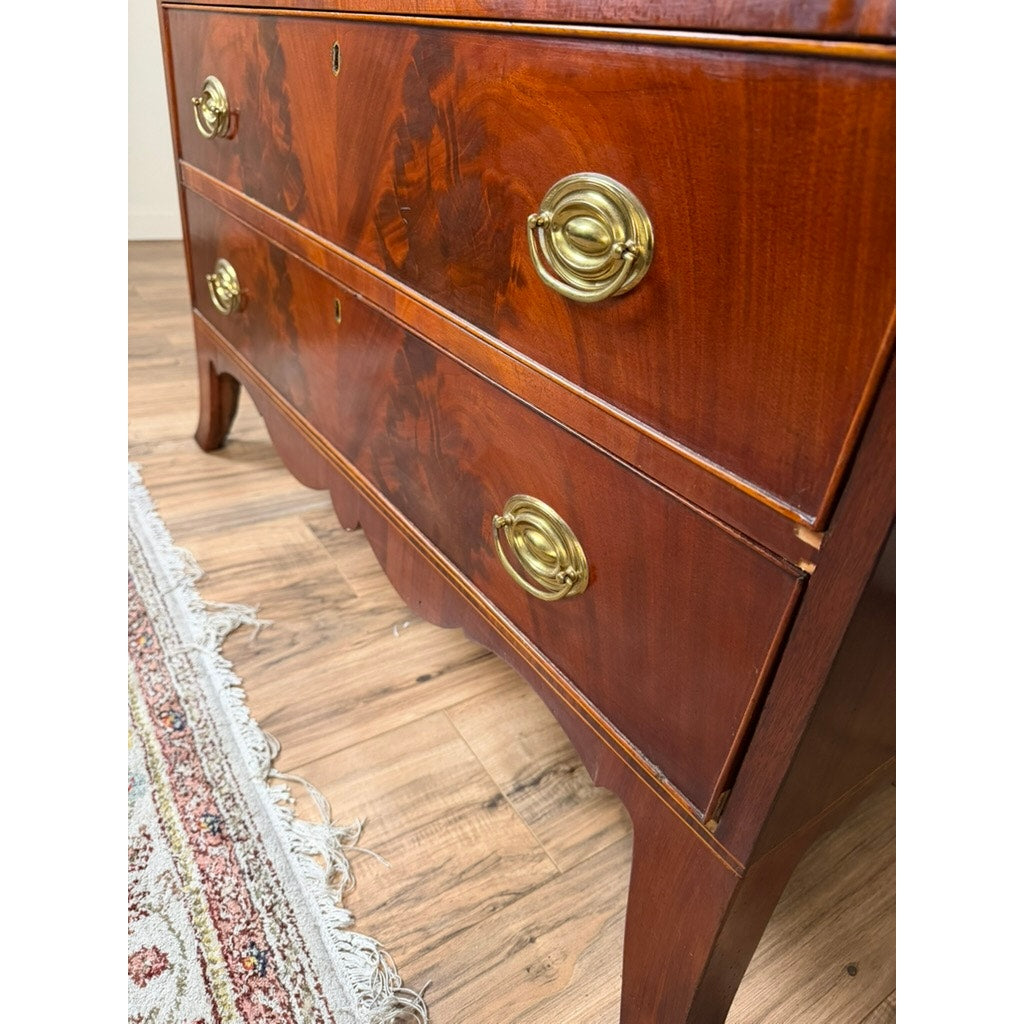 Close-up of a polished Federal Flame Mahogany Secretary Bookcase with Eagle Finial, c. 1810–1825, showing two drawers with brass handles. The bookcase, a testament to the refined craftsmanship of early 19th-century American cabinetmaking, sits on wooden flooring near a fringed patterned rug. The flame mahogany grain and fine finish are prominently visible.