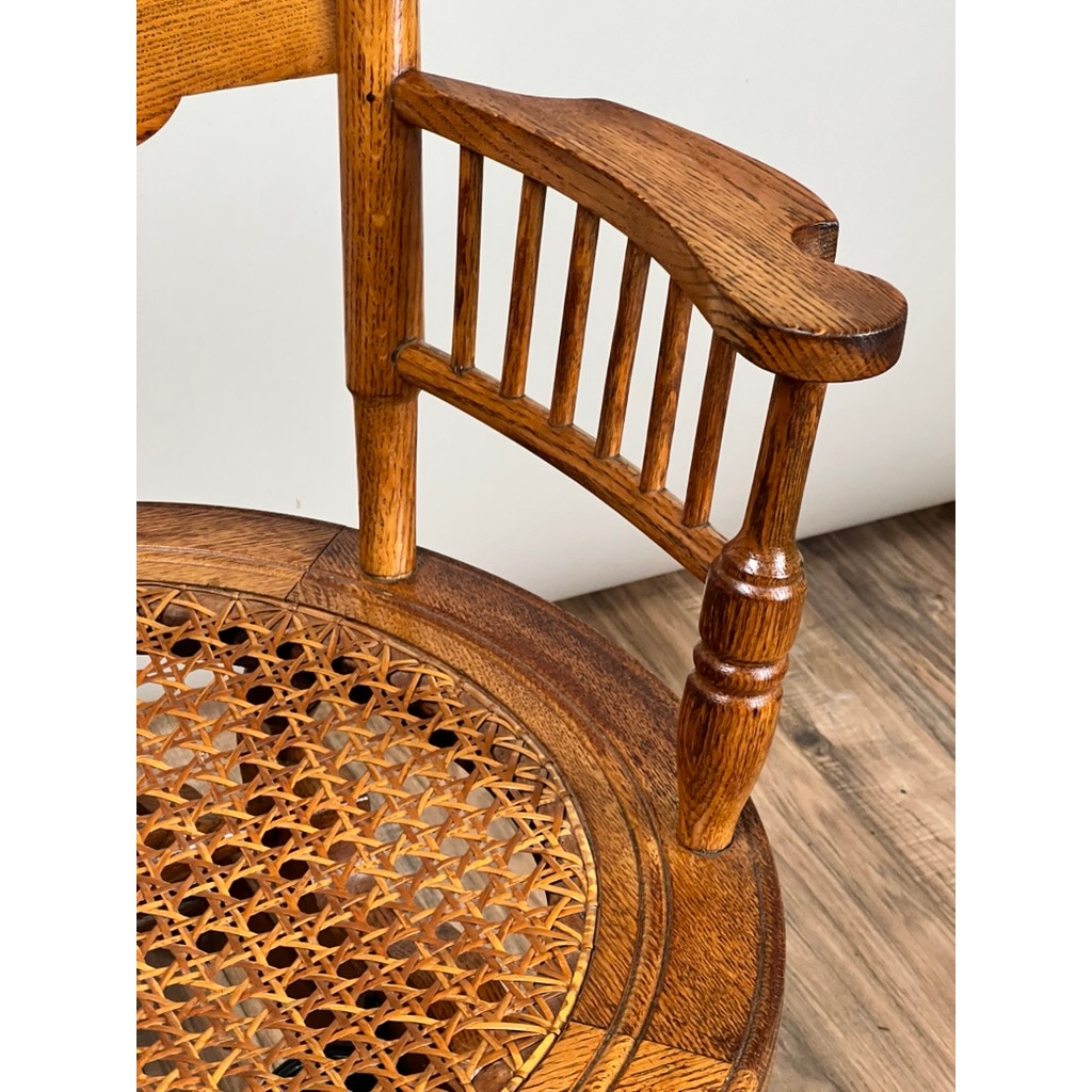Close-up of an Antique Victorian Oak Platform Rocker (c. Late 19th Century) with curved armrest, vertical spindles, and woven cane seat, shown on a wood-patterned floor against a plain light background. Brand: None/Unknown.