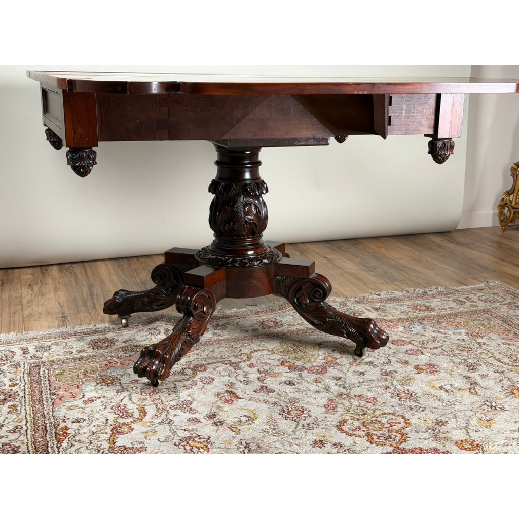 American Classical Carved Mahogany Drop-Leaf Breakfast Table, c. 1825–1840, featuring an ornate pedestal base with carved foliage and four carved feet with casters.