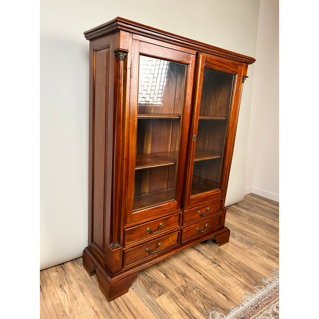 The Vintage Victorian Style Glass Door Mahogany Bookcase features two interior shelves, four lower drawers, decorative trim, and brass handles. It stands on a wooden floor against a light-colored wall. Brand: None and/or Unknown.