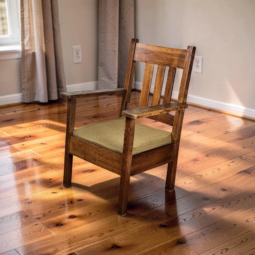 An Antique Quarter Sawn Mission Oak Arm Chair by Quaint Furniture c. 1920 with armrests and a green cushioned seat is placed on a polished hardwood floor in a sunlit room with beige walls, window, and light curtains.