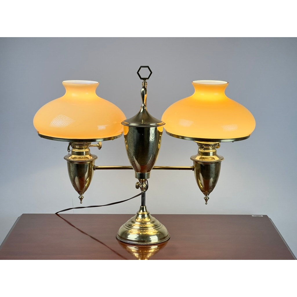 A Bradley and Hubbard antique brass double-arm student lamp with caramel shades rests on a polished wooden surface against a plain gray background.
