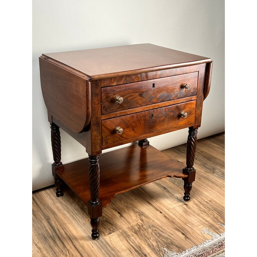 An Antique Federal Mahogany Two Drawer Work Stand c. 1820, featuring lion's head pulls, classic carved legs, a lower shelf, and drop-leaf sides, is shown on a wood floor against a plain wall. Brand: None/Unknown.