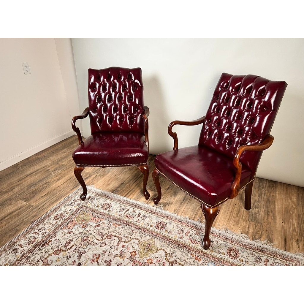 A pair of Chippendale Style Tufted Burgundy Leather Library Armchairs sit on a patterned rug in a room with wood flooring and light-colored walls, their tufted backs and wooden frames evoking timeless vintage elegance.