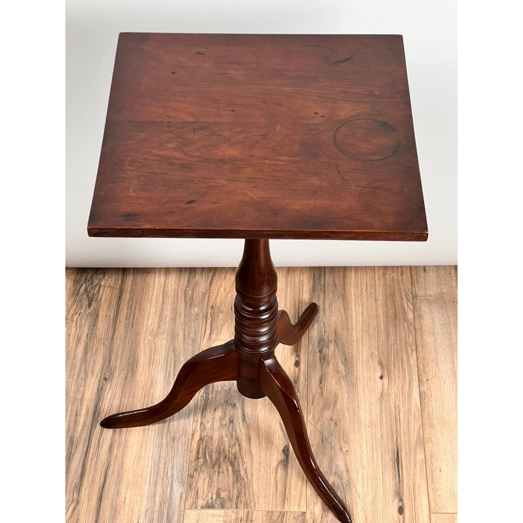 The Antique Low Cherry Candle Stand (c. Mid 19th Century) features a small square top, dark cherry finish, turned column, and three curved legs—an elegant early American piece perfect for display against a plain white wall. Brand unknown.