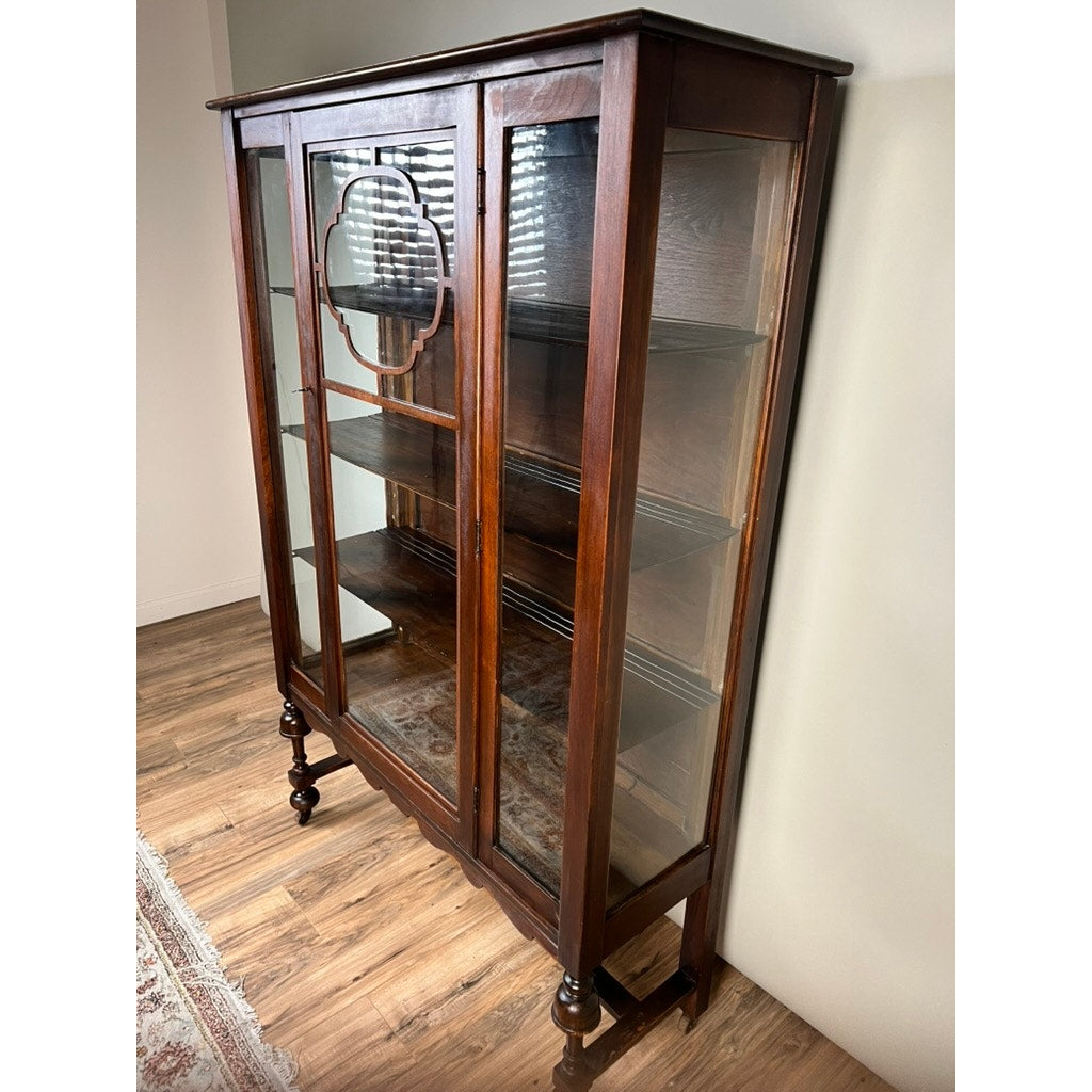 Depression Era mahogany curio display cabinet with glass sides, circa 1930s, featuring decorative wood trim and standing on turned legs—a striking vintage piece with shelves enclosed by glass doors—shown against a light-colored wall on a wooden floor.