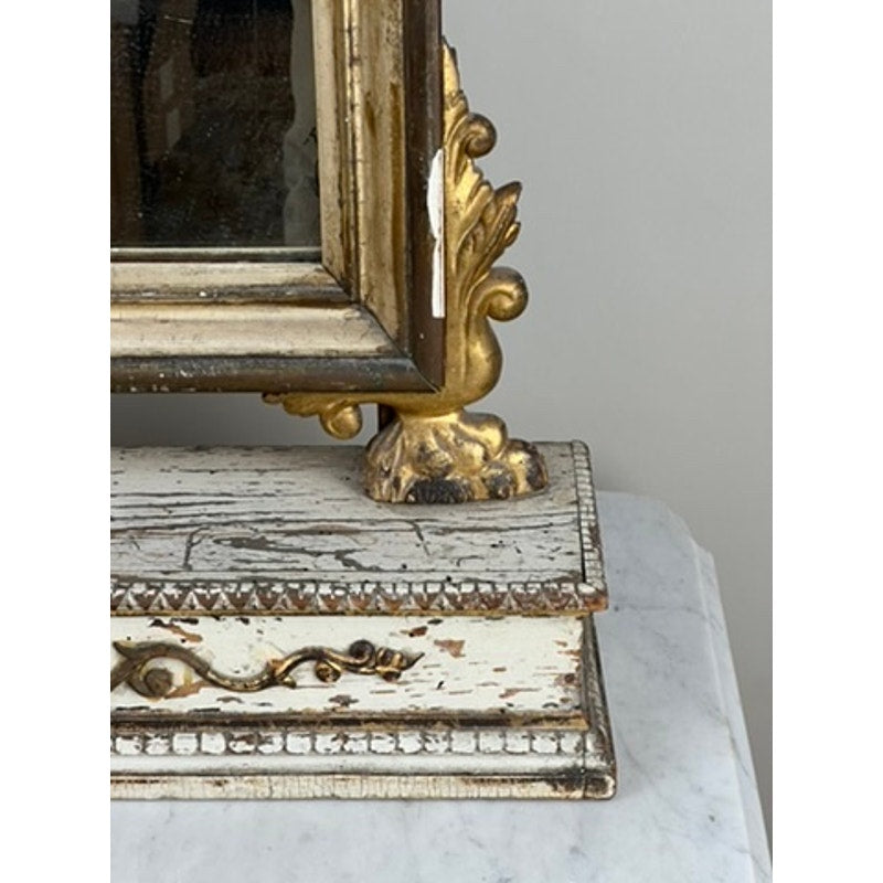 Close-up of an ornate gilt wood frame corner from an Antique French Empire Vanity or Table Mirror (mid-19th century), resting on a distressed, white-painted carved base atop a marble surface against a plain background.
