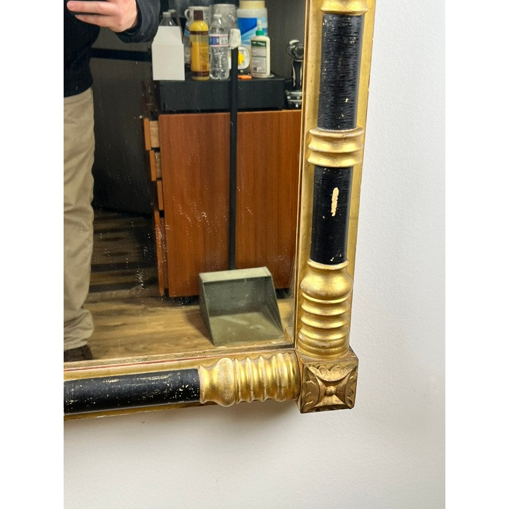 A partial mirror reflection shows a person’s lower body, an Antique Federal Period Giltwood Mirror with Rosette Corners and Cornucopia c. 1830, a dustpan on the floor, and shelves with cleaning supplies in the background.