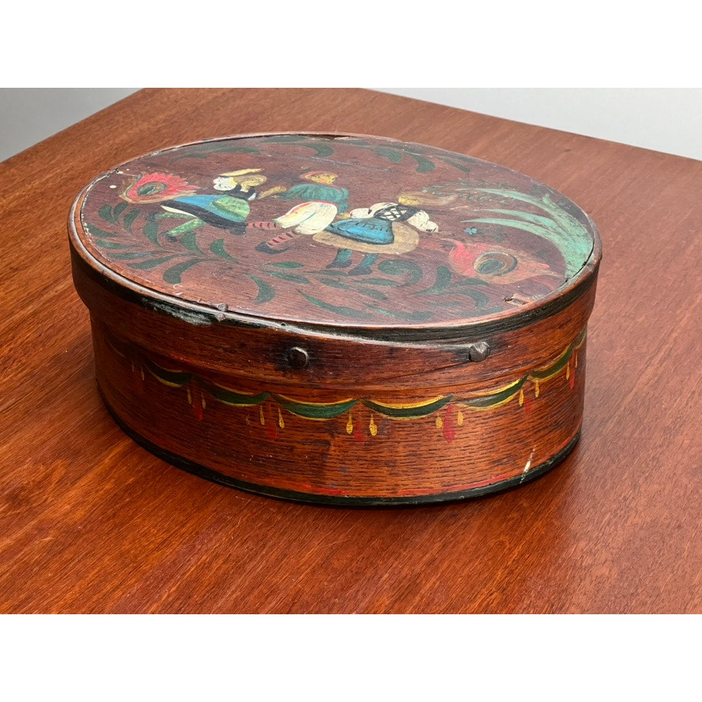 An Antique German Polychrome Painted Bentwood "Spanschachtel" Ribbon Box, c. late 18th century, features colorful European folk art floral and figure designs on an oval-shaped body with a hinged lid, displayed on a reddish-brown surface.