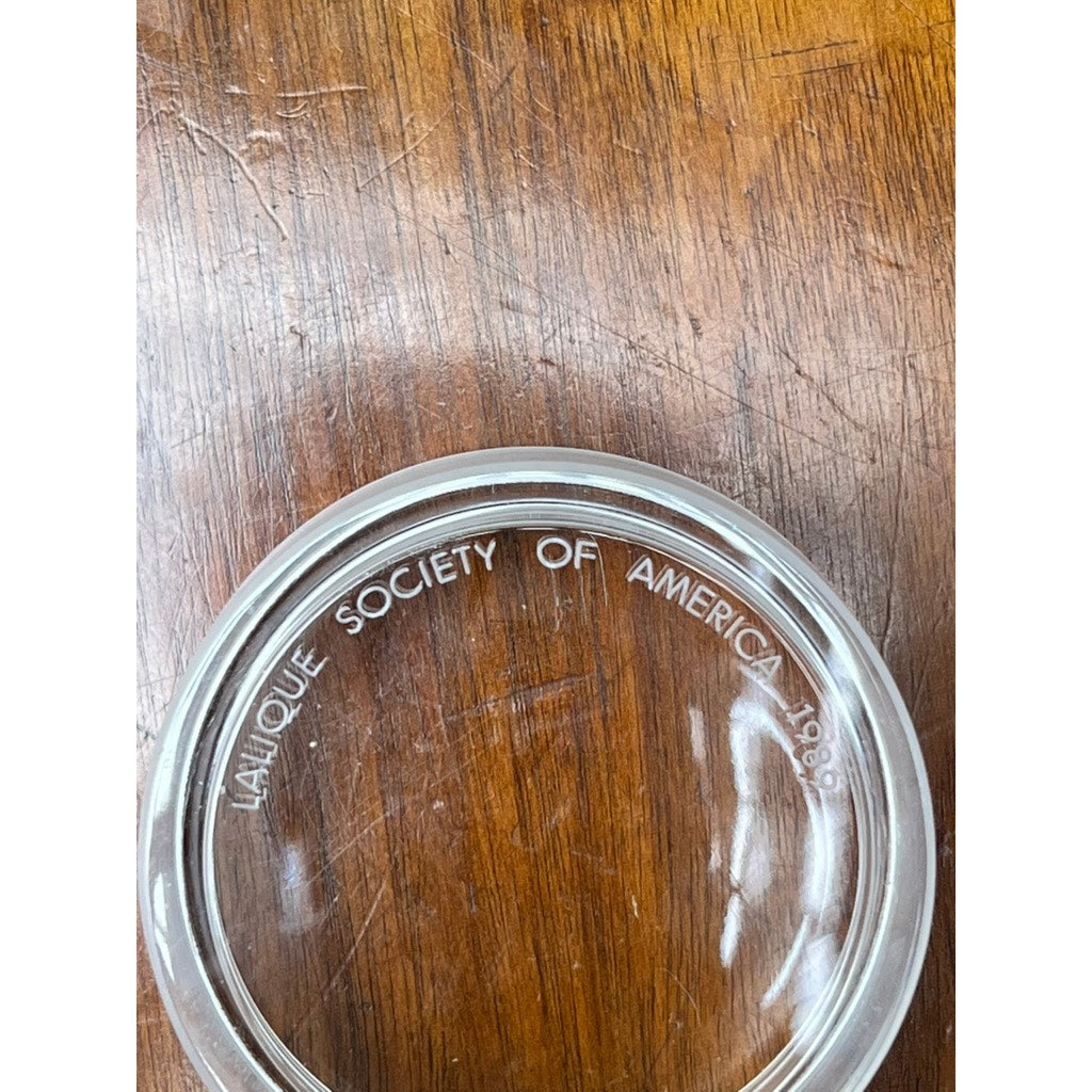 A Vintage Lalique France Ballerina Frosted Crystal Covered Powder Box by Lalique, engraved with "LALIQUE SOCIETY OF AMERICA 1989" along the edge, rests on a wooden surface.