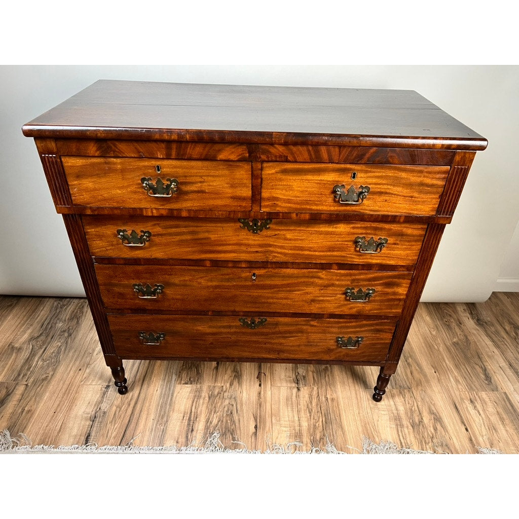 An Antique George III Sheraton Flame Mahogany Chest of Drawers c. Early 19th Century, with two small top drawers, three larger below, metal handles, polished finish, and four carved legs, stands on a wooden floor.
