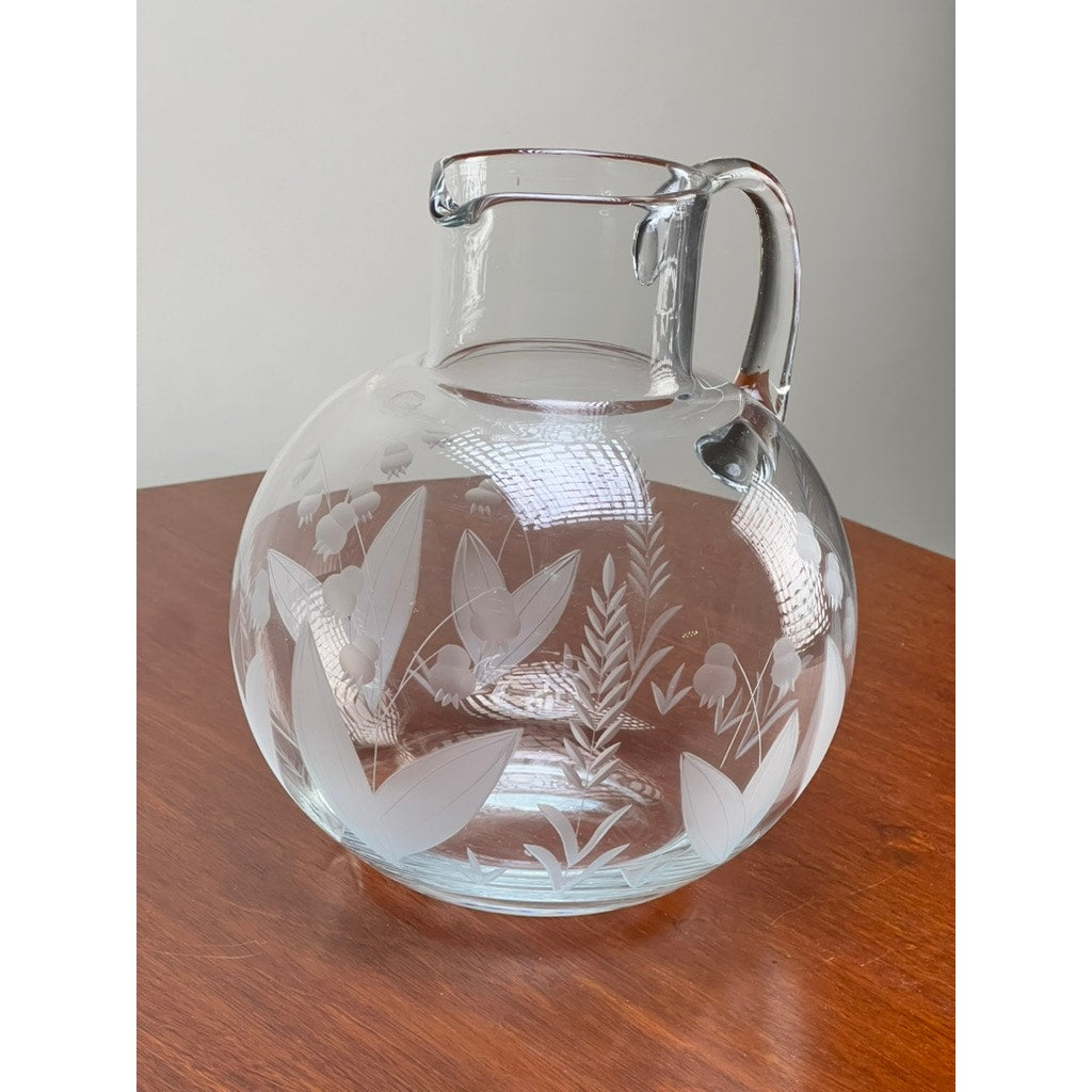A Vintage Tiffany & Co. Etched Crystal Pitcher featuring a lily of the valley floral motif rests on a wooden surface, its elegant wide body and handle highlighted against a plain light gray background.