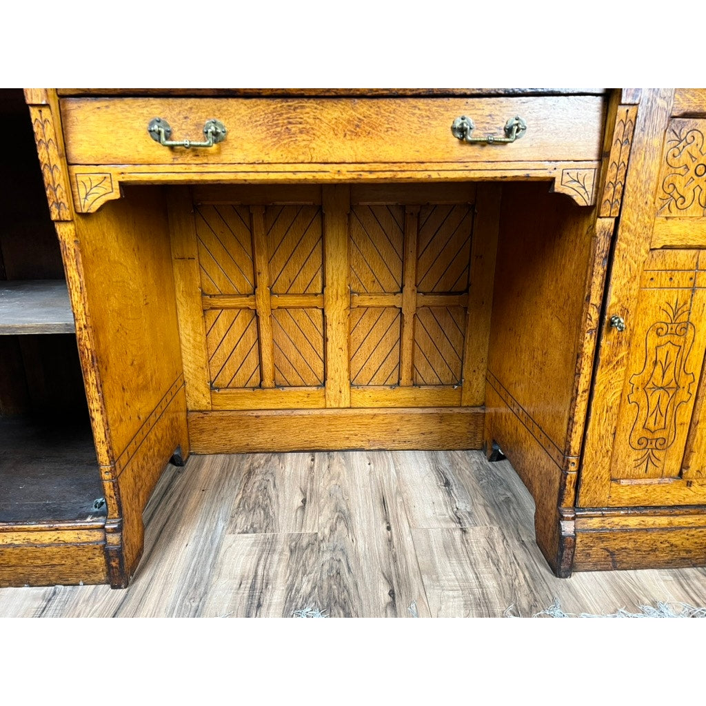 Close-up of an Antique Victorian Aesthetic Movement Oak Desk (late 19th c.) in original finish, showing wood grain, brass handles, carved details, empty shelves, and central recessed panel on a wood-patterned floor. Brand: unknown.